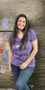 Codi Leary Leitch wearing a purple shirt and jeans leaning against a rock wall with painted handprints on it